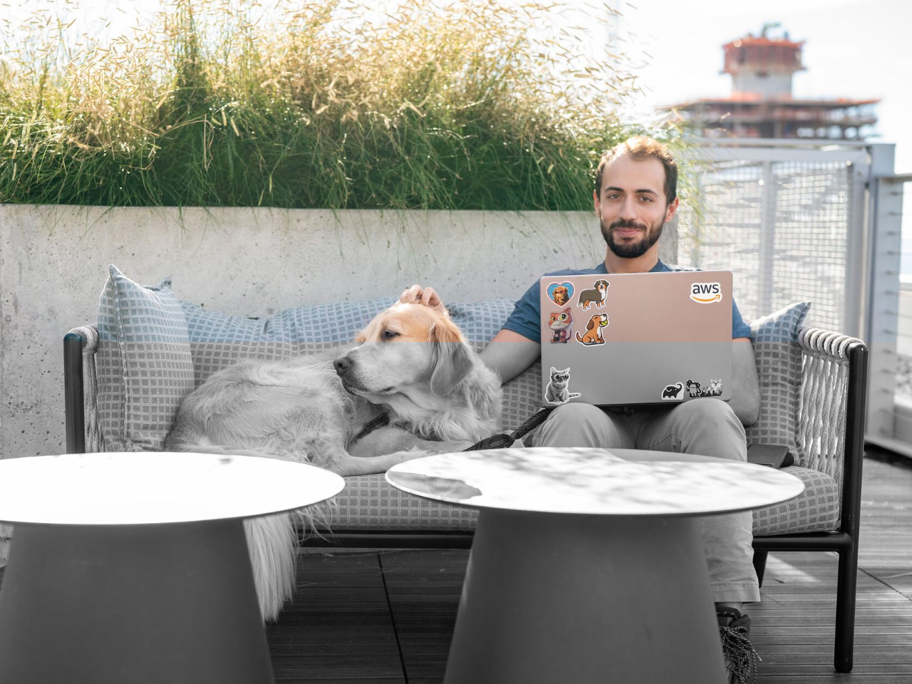 Ein Bild eines Mannes, der an einem Tisch im Freien in einem Amazon-B&uuml;ro sitzt, mit seinem Laptop vor sich und einem gro&szlig;en gelben Hund an seiner Seite. Der Mann streichelt den Hund und schaut in die Kamera.