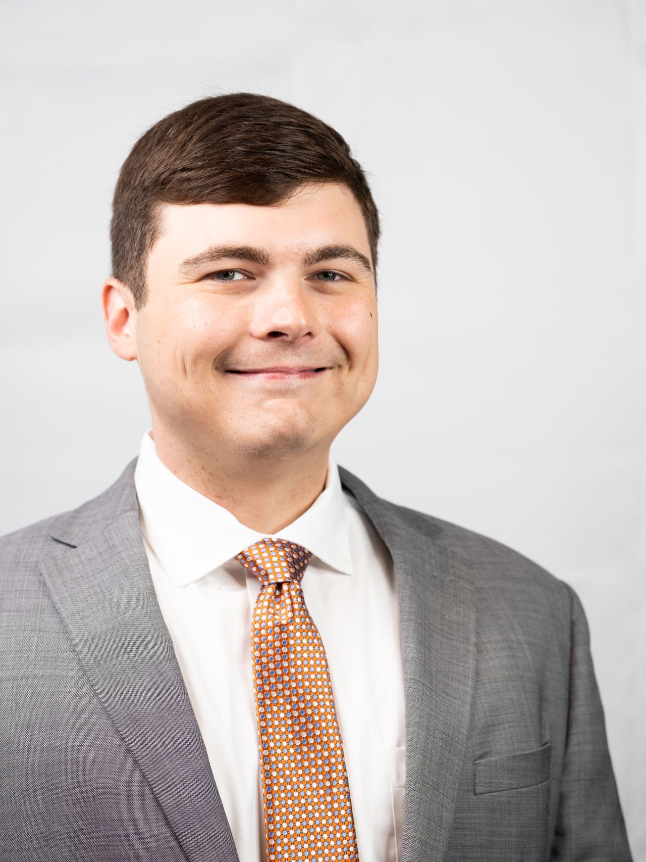 Portrait of a businessman wearing a grey suit, white shirt, and orange patterned tie, smiling against a light background.