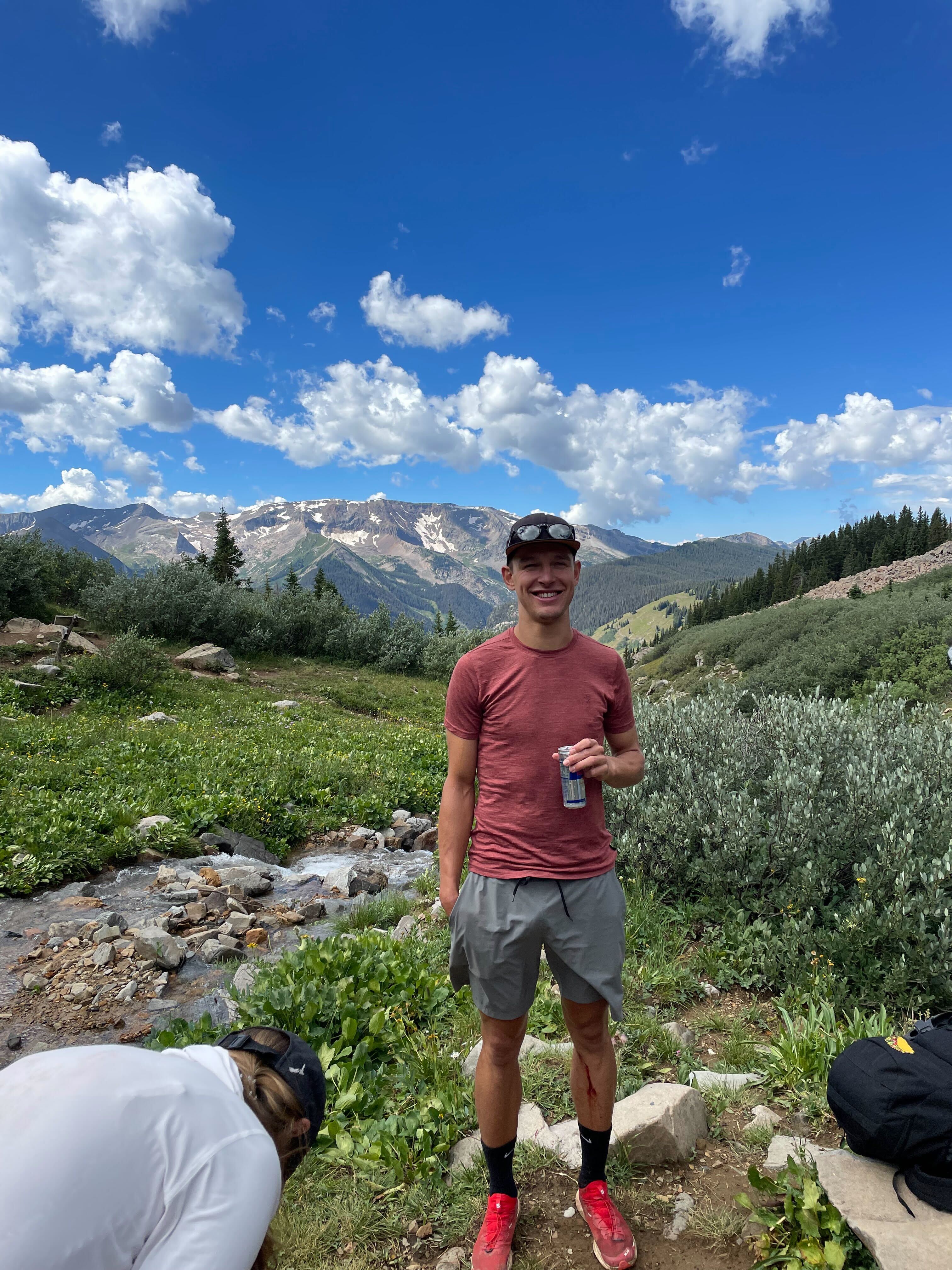 A man standing and smiling beside a mountain stream on a sunny day, with alpine mountains and blue sky with clouds in the background. He is wearing a red shirt, gray shorts, red shoes, and a cap, holding a drink, surrounded by lush greenery.