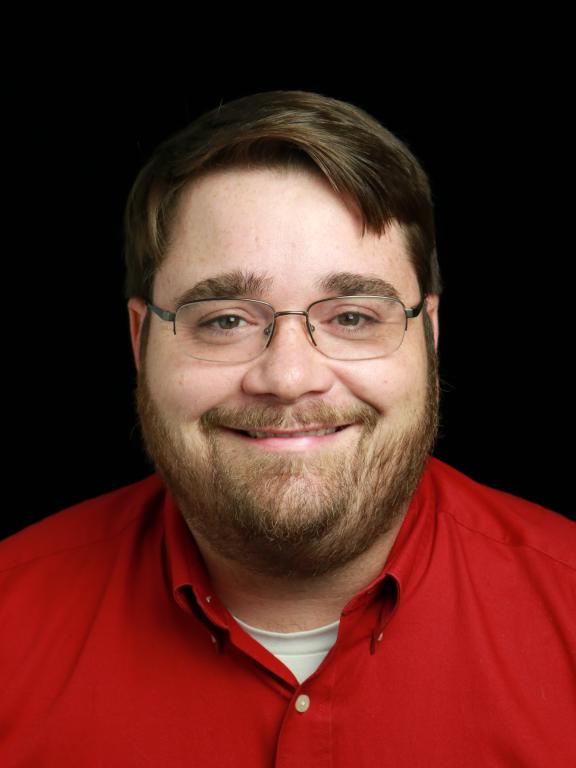 A portrait photograph of a man with brown hair and a beard, wearing glasses and a red shirt, smiling against a black background.