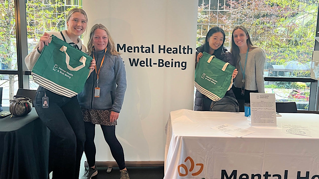 Four people smile and stand behind a table displaying 'Mental Health & Well-Being' at an Amazon affinity group event. Two of them hold tote bags with the Mental Health & Well-Being logo. The event appears to promote awareness and support for mental health and well-being among employees.