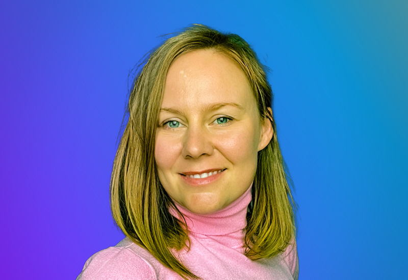 A person with shoulder-length blonde hair wearing a pink turtleneck against a gradient blue and purple background.