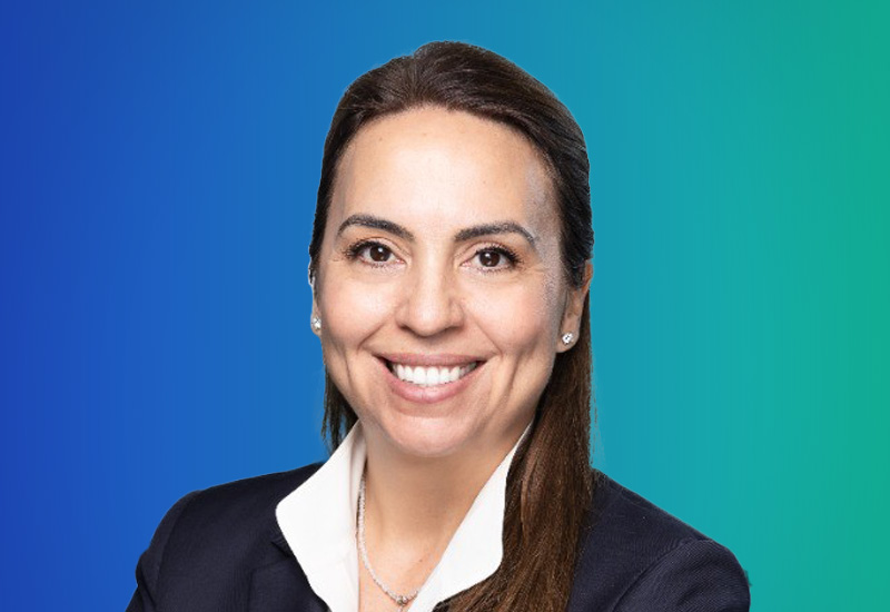 A professional woman smiling, wearing a white shirt and dark blazer, posed in front of a blue to green gradient background.