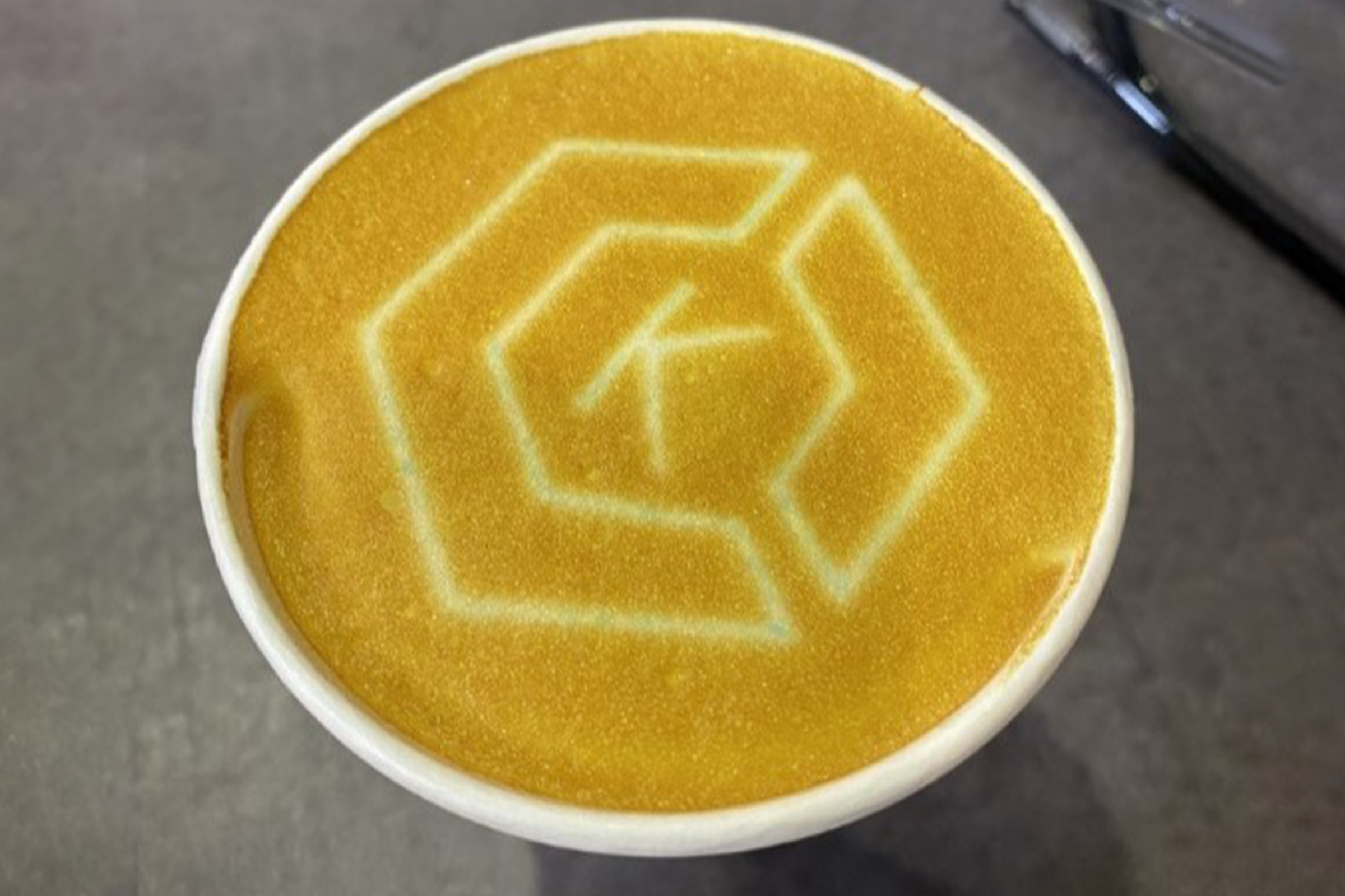 A coffee latte with KubeCon branding in the foam, presented at an AWS Retail event. The cup's surface features a geometric pattern associated with containers or Kubernetes, symbolizing the cloud native ecosystem, likely photographed at a technology conference or event.