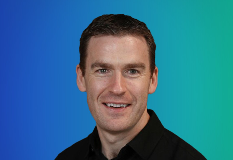 Photograph of a smiling person with short dark hair, wearing a black shirt, in front of a blue to green gradient background.