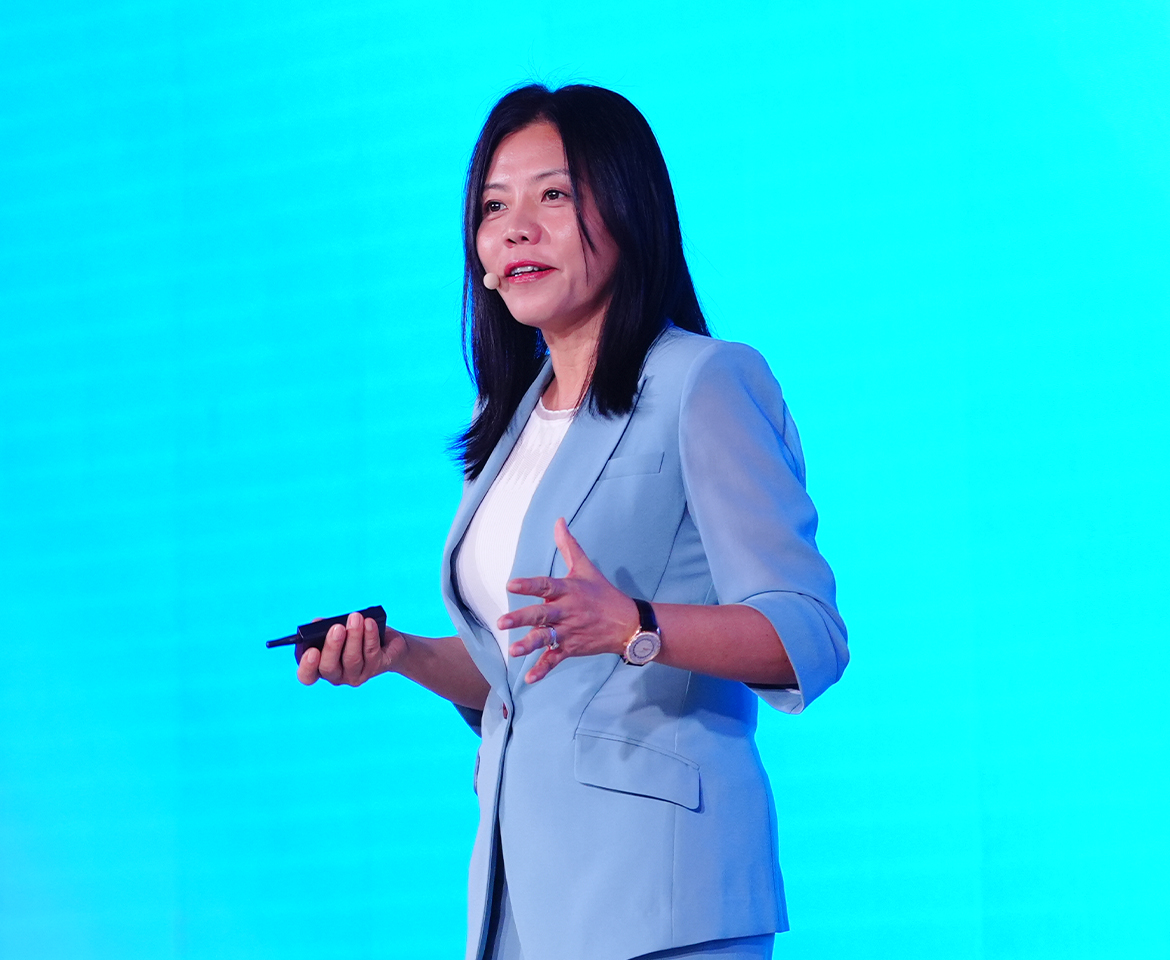 A woman in a light blue suit giving a presentation against a bright blue background, holding a clicker in one hand.