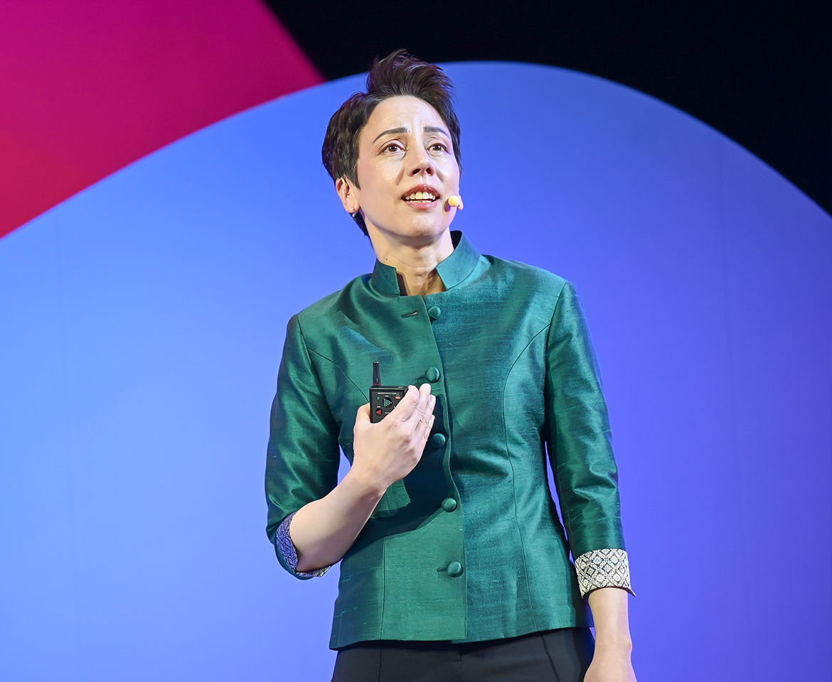 Person wearing a green buttoned jacket holding a microphone device against a colorful backdrop.