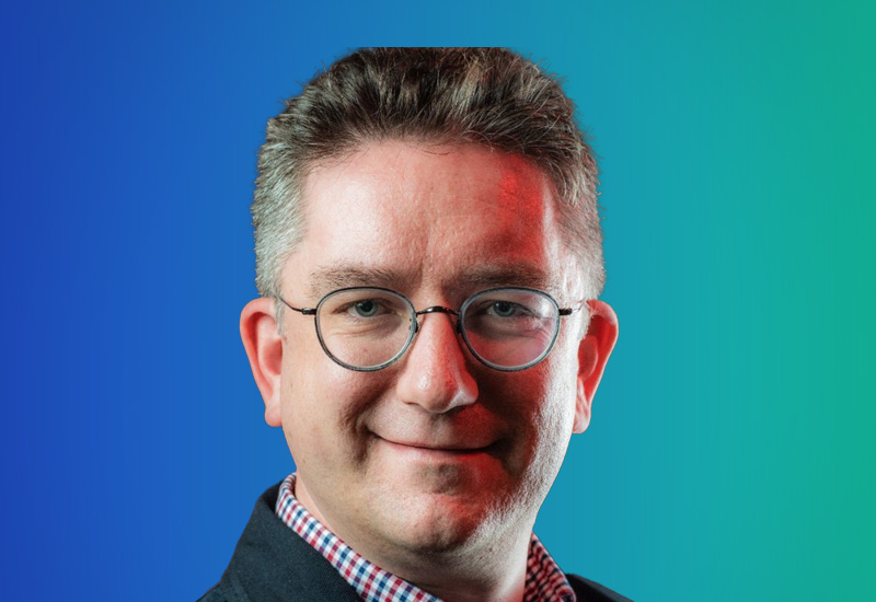 A man with short hair and glasses, wearing a suit and checkered shirt, is smiling in front of a blue and green gradient background.