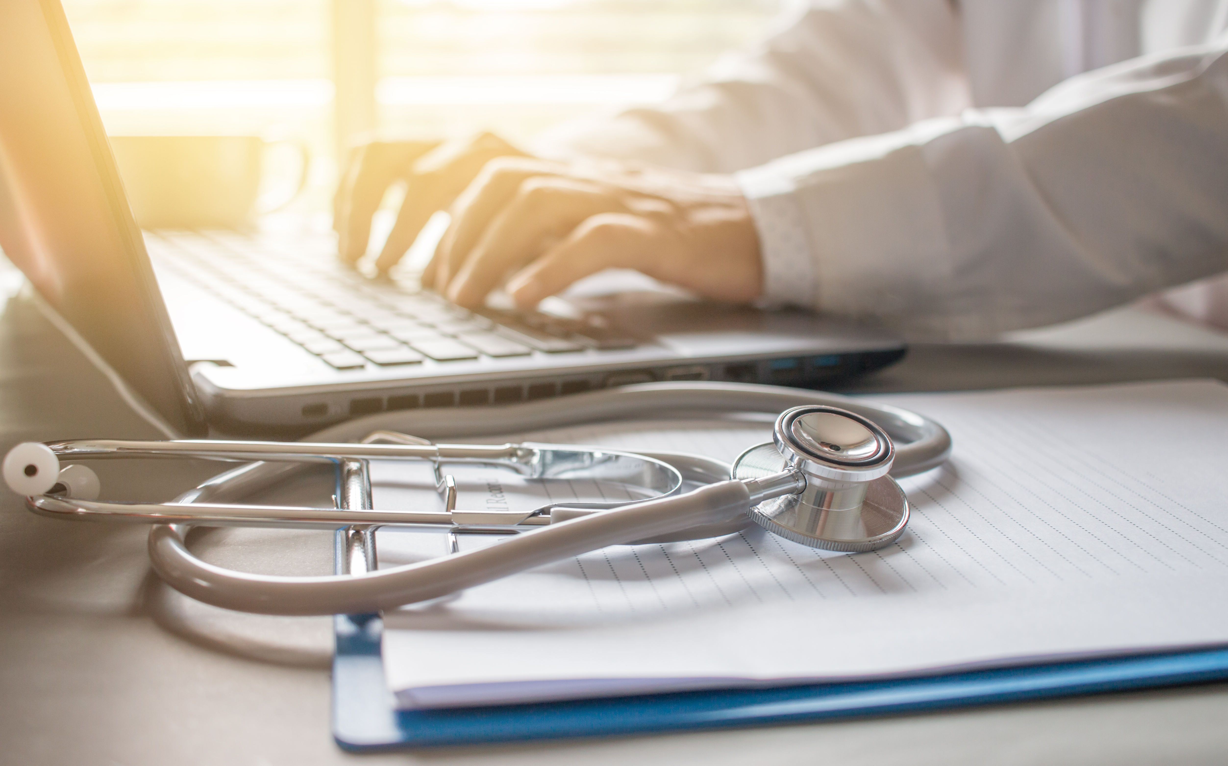 A close-up of a doctor typing on a laptop with a stethoscope and prescription clipboard on the desk in a hospital setting.