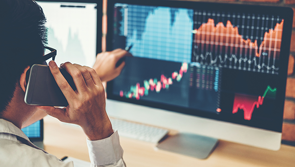 A person analyzing stock market data on computer monitors, showing various charts and graphs. The individual is speaking on the phone while pointing at financial data, representing financial analysis or stock trading activities.