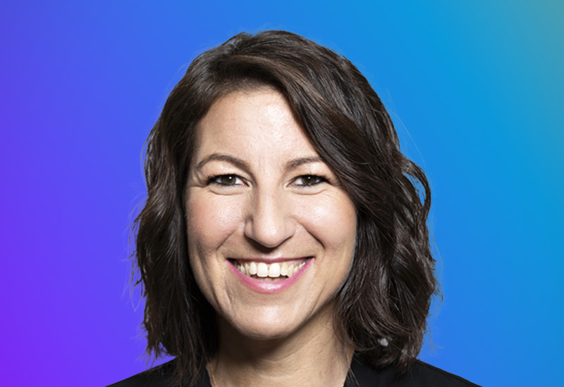 A professional portrait of a smiling woman with shoulder-length wavy brown hair, set against a blue and purple gradient background.