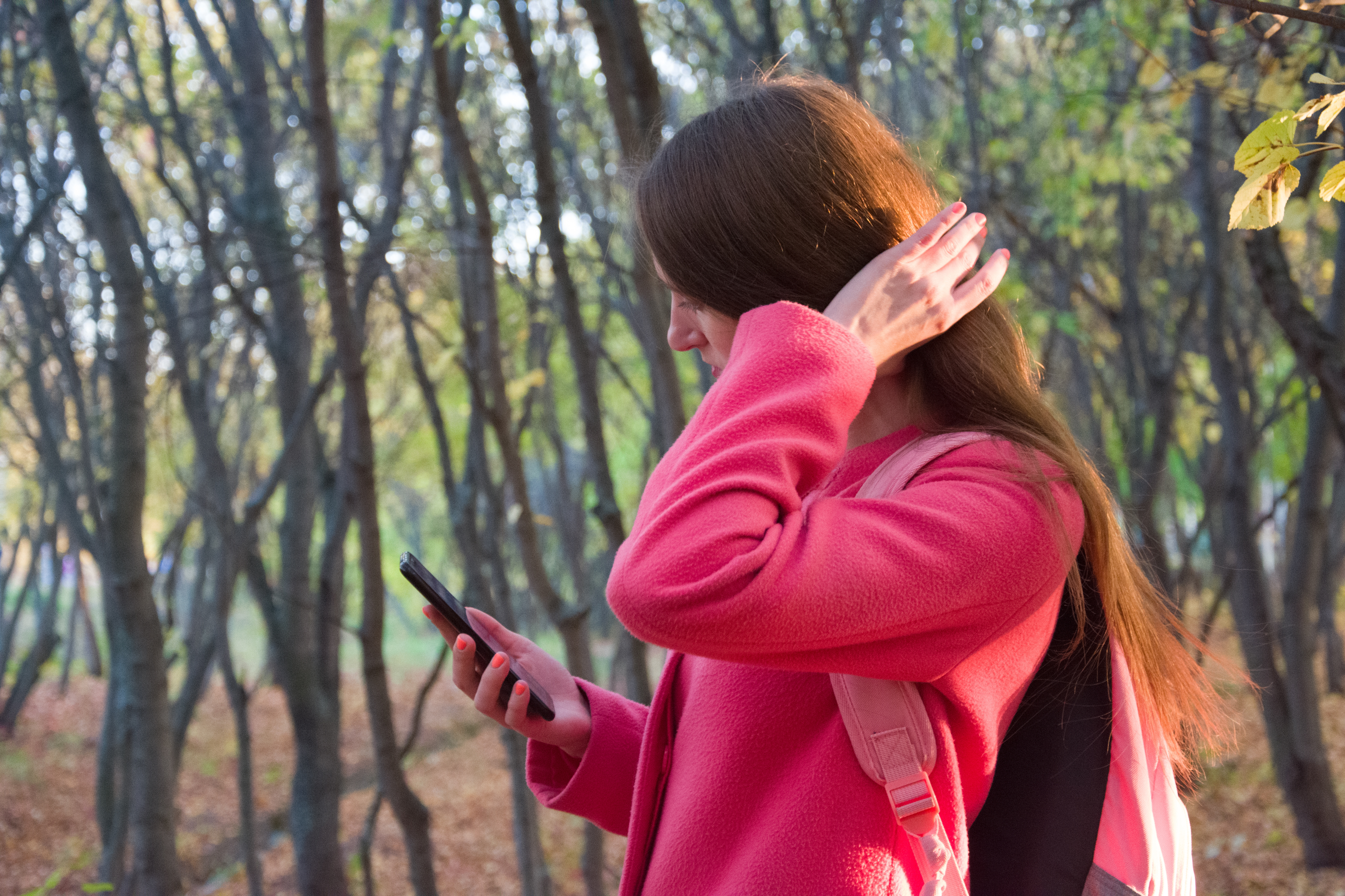 Side view of unrecognizable young female with long straight sandy hair surfing internet in autumn park in sunlight on blurred background