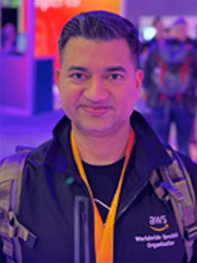 A person wearing an AWS jacket with an event badge and backpack, standing in an indoor event or conference setting, with people and signage visible in the background.