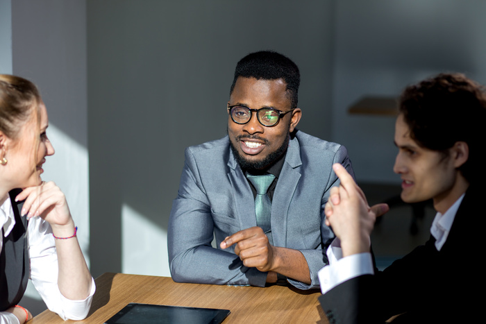 Three people in formal attire sitting at a table in a well-lit office setting, engaged in discussion.