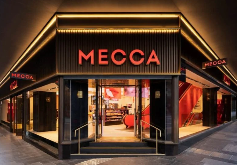 The entrance of a Mecca retail store, showcasing the illuminated MECCA brand signage and a modern, stylish storefront design with visible displays inside.