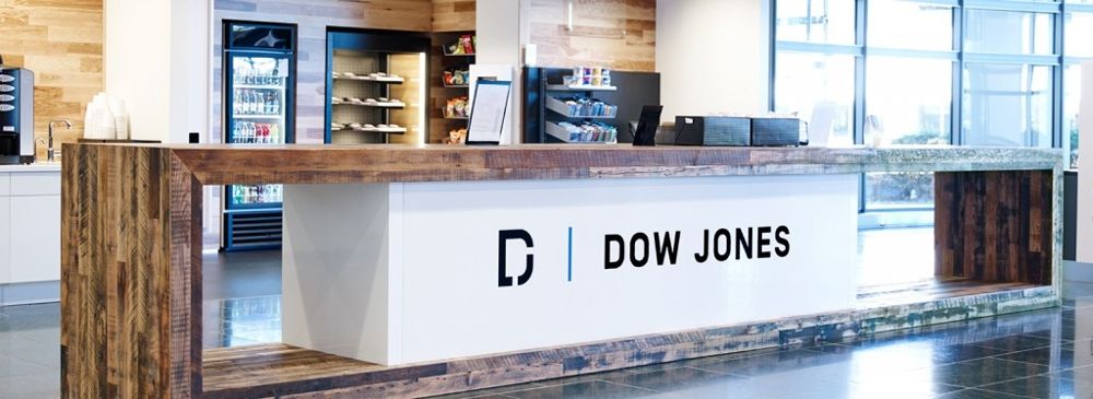 A modern office reception area with a wooden counter displaying the Dow Jones logo. The background features shelves and refrigerators with snacks and drinks, situated in a bright, open space with large windows.