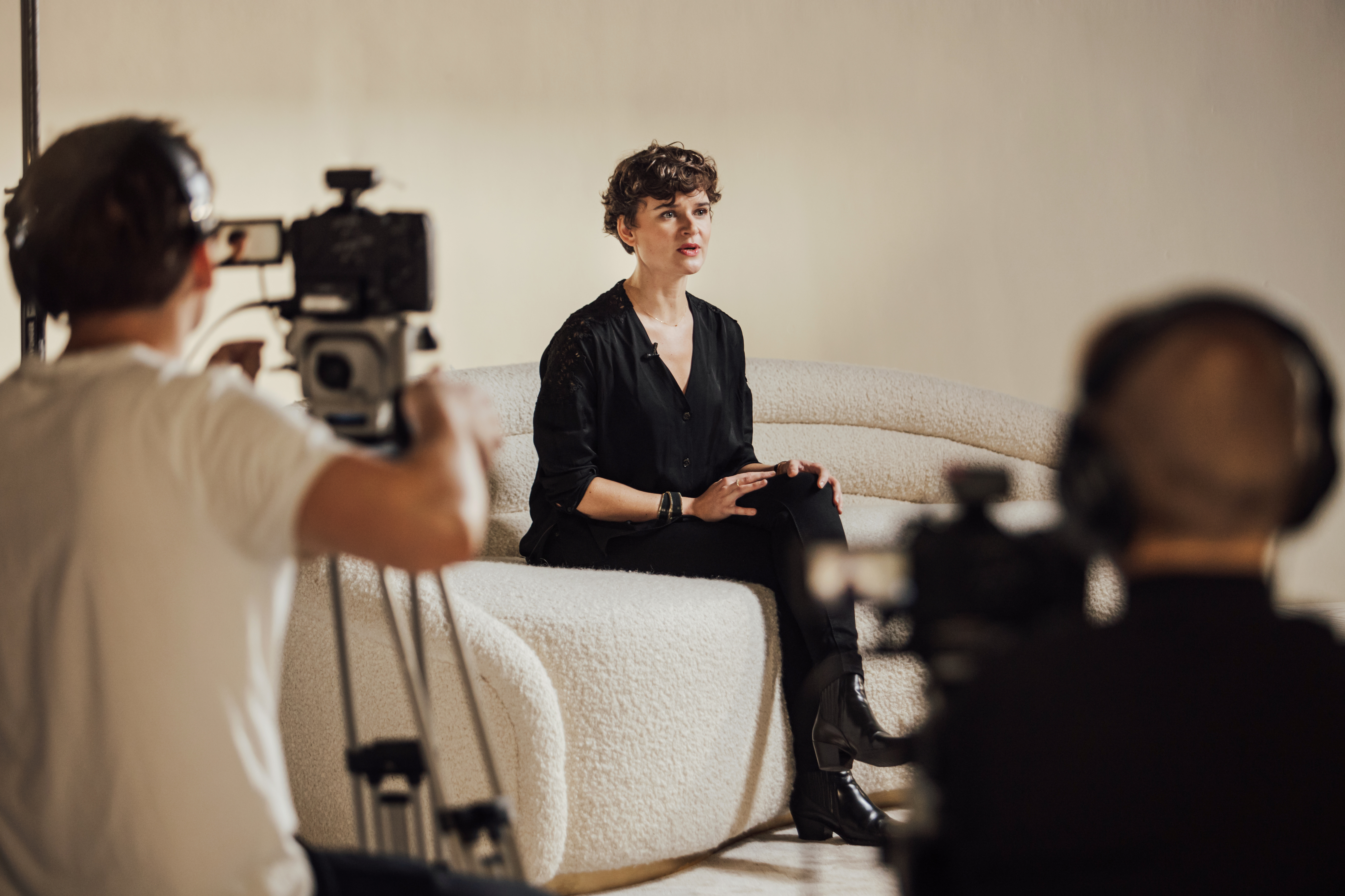 A person dressed in black sits on a textured white couch during a video recording session, with cameras and crew visible in the foreground.