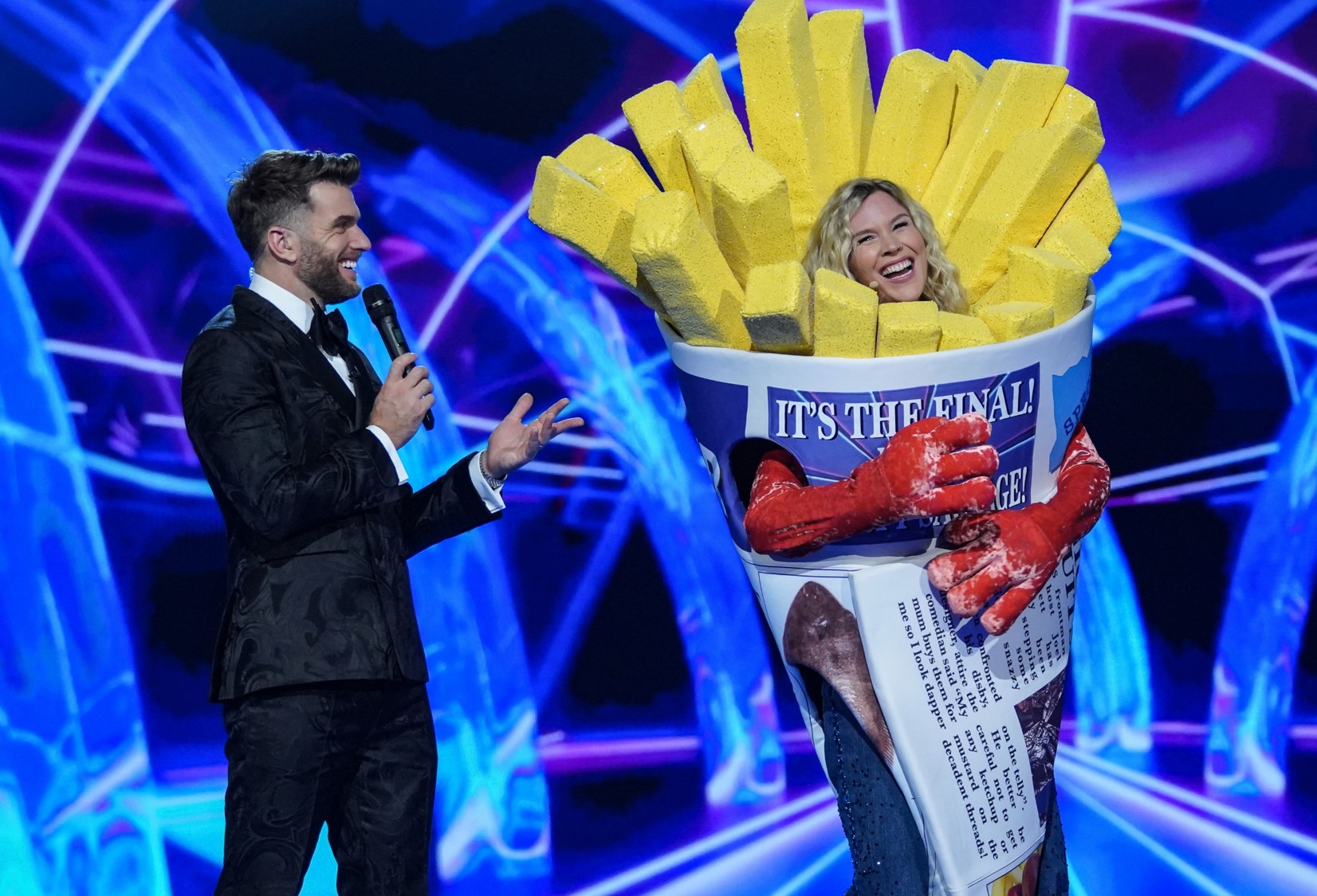 A man in a suit and bow tie interviews a smiling woman dressed in a humorous French fries costume on a brightly lit stage with a blue and purple background.