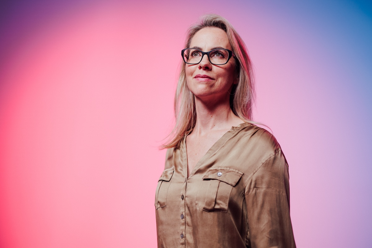 Portrait of a woman wearing glasses and a beige blouse, standing in front of a pink and blue gradient background.