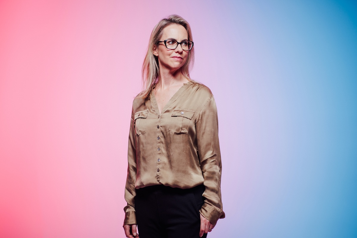 A woman wearing glasses and a satin blouse stands against a pink and blue gradient background, looking to the side.