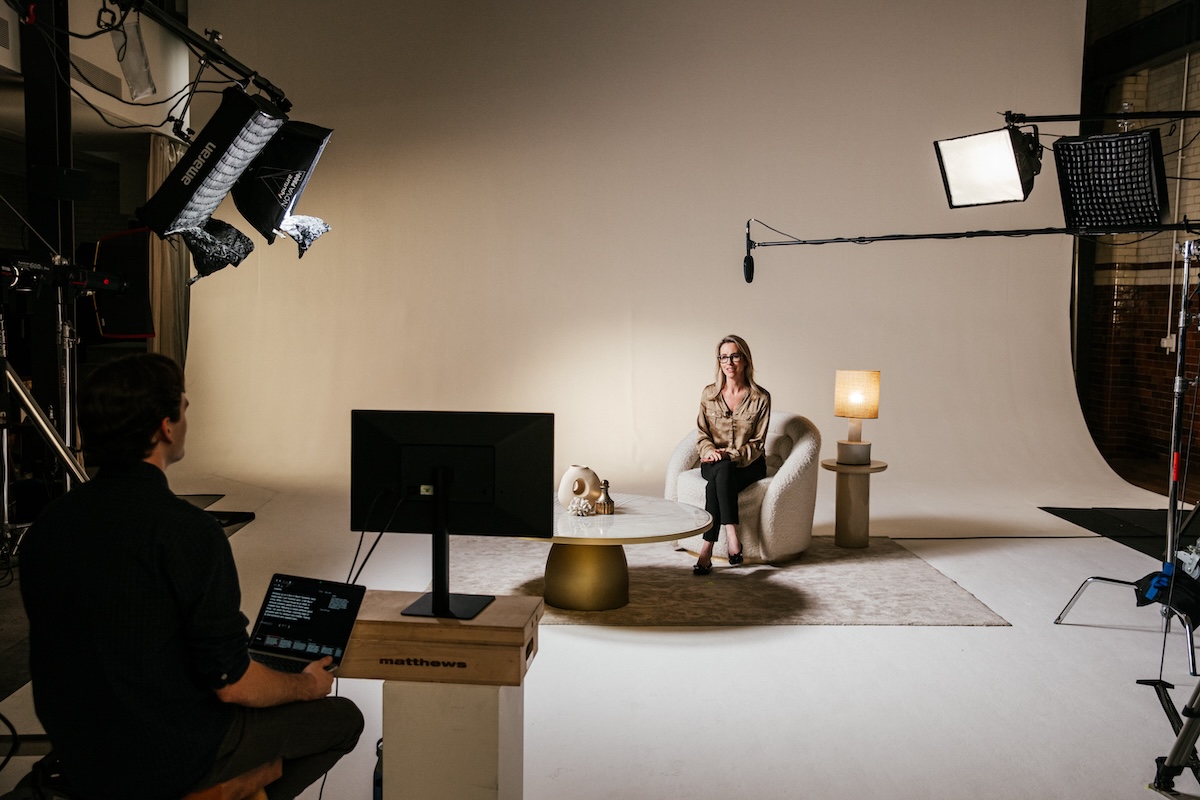 A behind-the-scenes view of a professional interview setup in a studio. Studio lights, camera equipment, and a crew member operating a laptop are visible as a person sits on a chair prepared for the interview under lighting and a boom microphone.