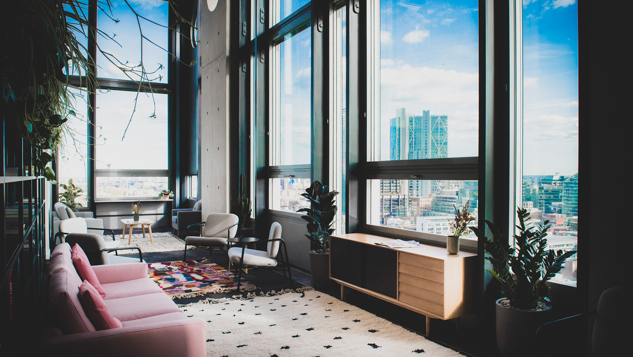 A stylish modern lounge area with large windows overlooking a cityscape, featuring comfortable seating, plants, and contemporary decor in natural light.