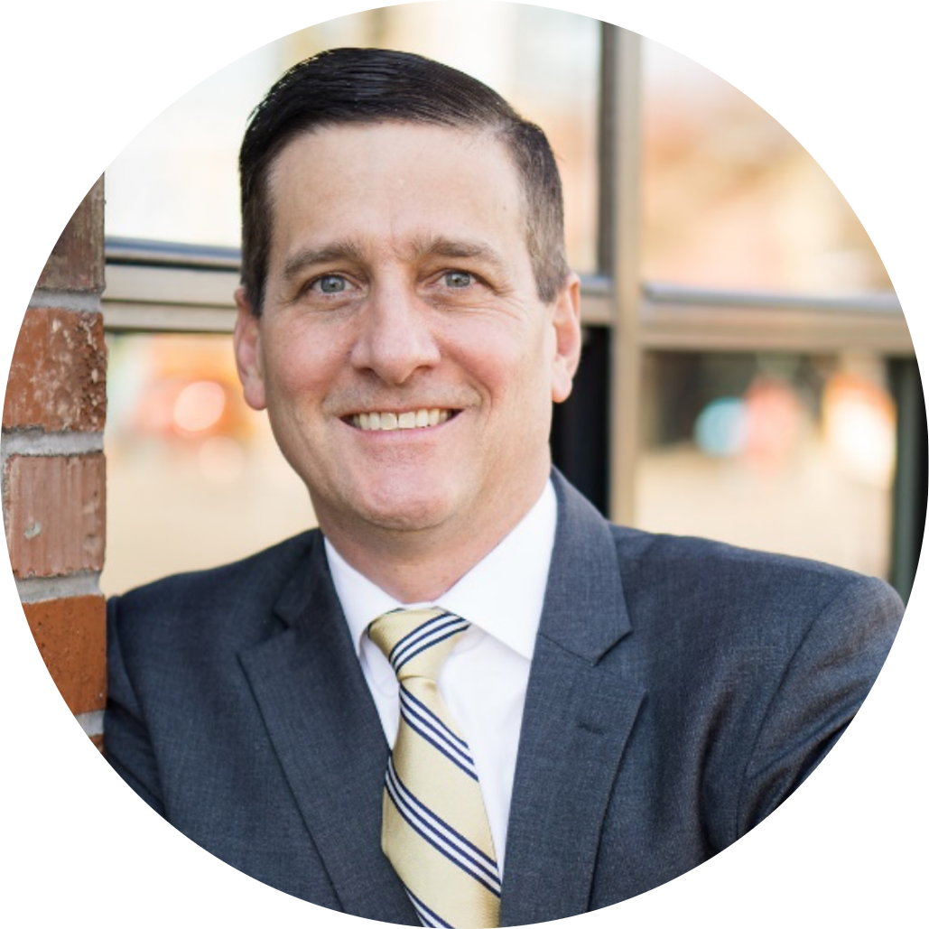 Professional business portrait of a man wearing a dark suit, dress shirt, and striped tie, smiling and standing near a brick wall and window.