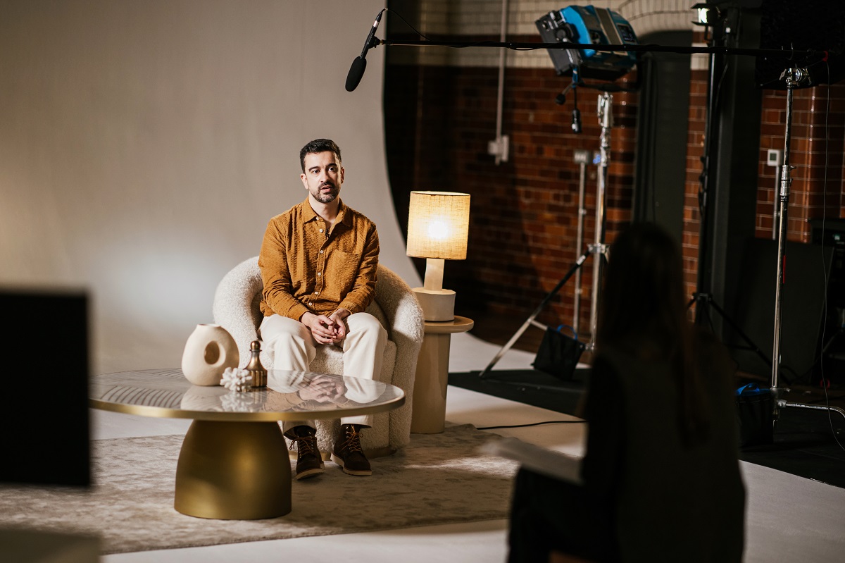 A man sits on an armchair in a studio setting, with a microphone, lighting equipment, and a camera visible. There is a table with decorative items and a lamp next to him, indicating a professional interview or video recording environment.