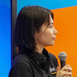 A person holding a microphone speaking at an AWS event, wearing an AWS-branded shirt, with a colorful background.