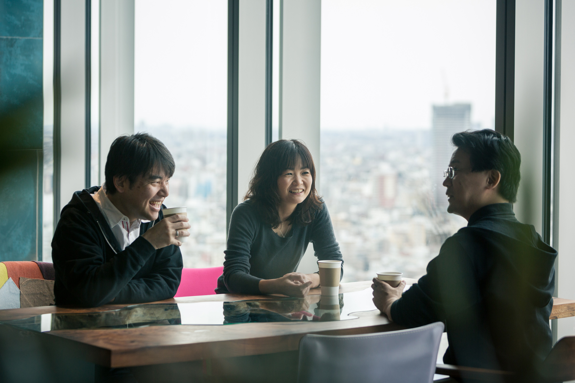Three people having a friendly conversation and enjoying coffee together in a modern office lounge with large windows and a cityscape view in the background.