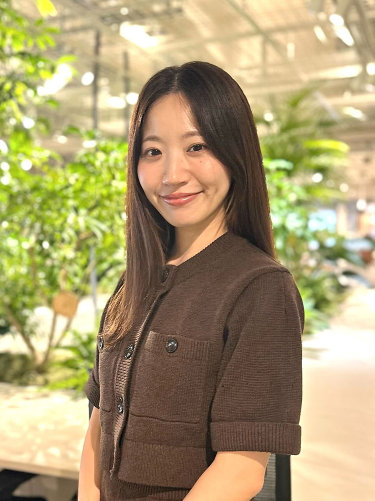 A woman wearing a brown top smiles while standing indoors with lush green plants in the background.