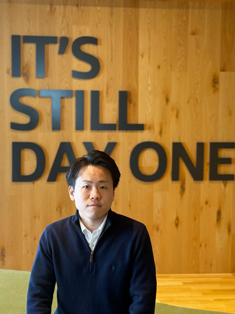A man is standing in front of a wooden wall with the bold text 'IT'S STILL DAY ONE' at an AWS office.