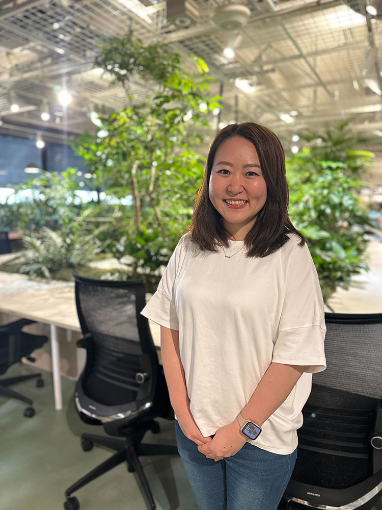 A woman standing and smiling in a modern office with abundant greenery and natural lighting, with office chairs and desks visible in the background.