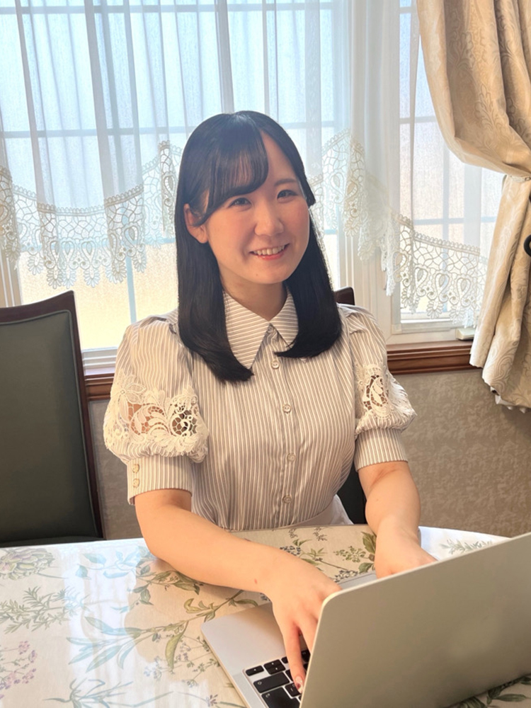 A woman smiling while using a laptop at a table, seated indoors with curtains and a window in the background.