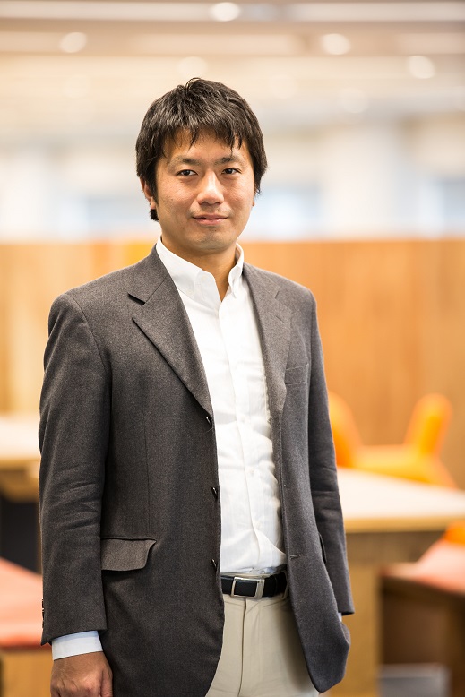 A professional portrait of a man in business attire, standing in a modern office setting with blurred desks and orange chairs in the background.