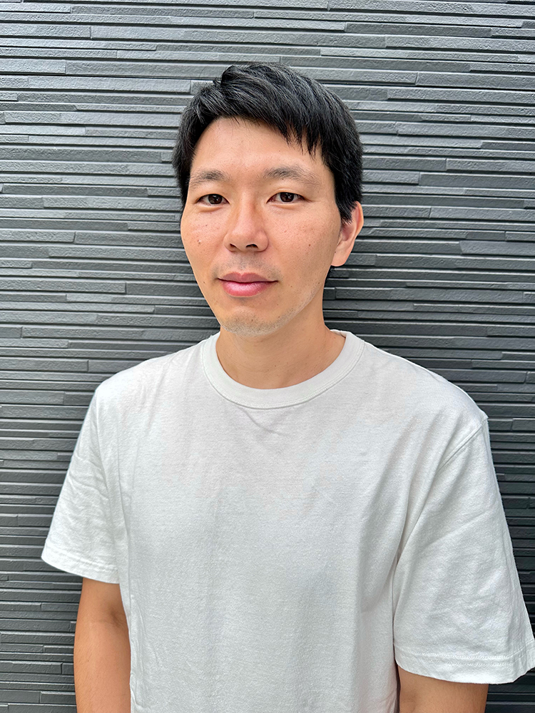 A man wearing a white t-shirt stands in front of a gray textured wall.