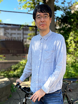 A man wearing glasses and a light blue shirt stands outdoors next to a black bicycle, with greenery and an urban background visible.