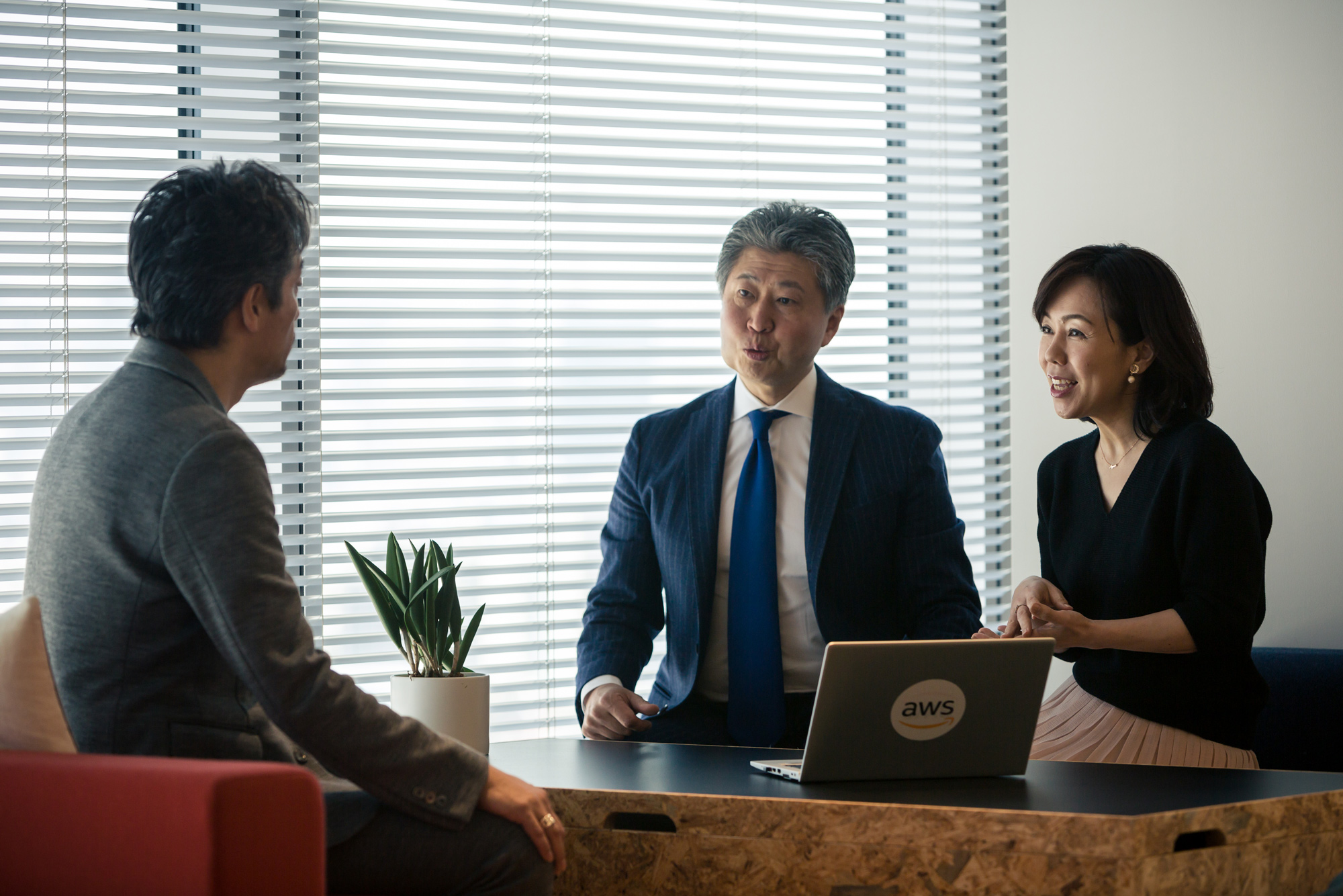 Three professionals having a business discussion at a table in a modern office, with an AWS-branded laptop visible on the desk.
