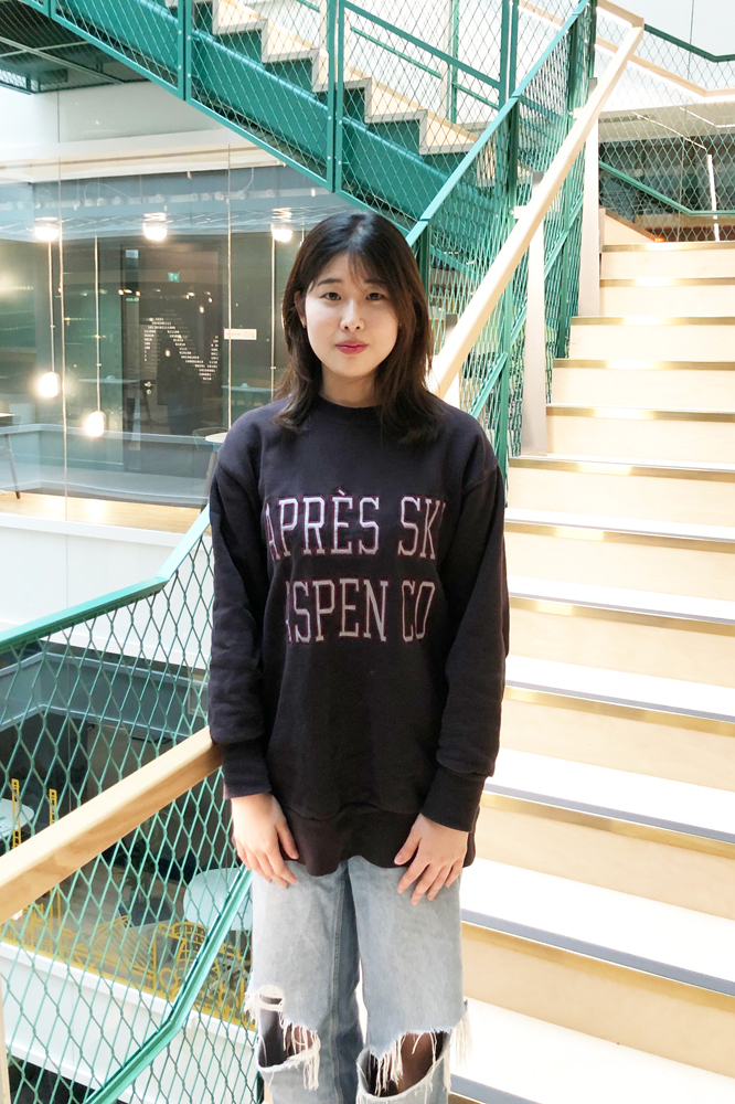 A woman stands on a modern staircase indoors, wearing a dark Après Ski Aspen CO sweatshirt and ripped jeans.