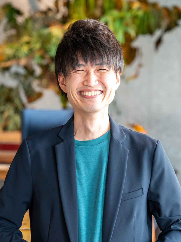 A man smiling brightly while wearing a teal shirt and a navy casual blazer, photographed indoors with a blurred background of greenery and wall art.