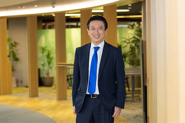A businessman in a dark suit and blue tie is smiling while standing in a modern office environment with wooden accents and green plants in the background.