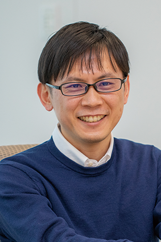 Portrait of a professional male DENSO employee wearing glasses and a navy sweater, smiling in an office setting.
