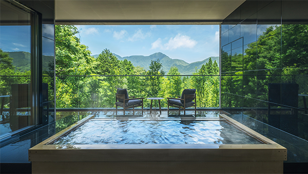 Interior view of an onsen hot spring room at Hoshino Resorts, featuring a panoramic window with a scenic view of mountains and lush green forest. The modern room includes a large soaking bath and two chairs by a table overlooking the natural landscape.