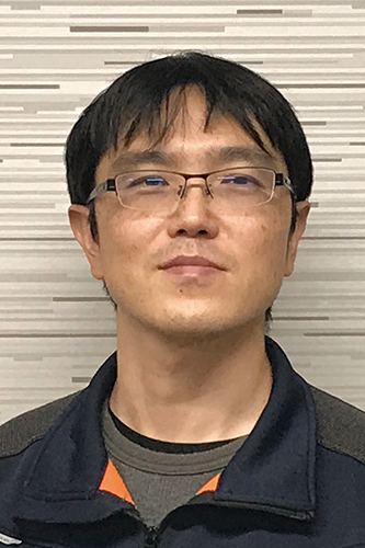 Portrait of a man in glasses with short black hair and casual attire, standing in front of a striped background.