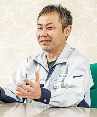A man in a Nabtesco uniform speaking at a table during an interview or case study discussion.
