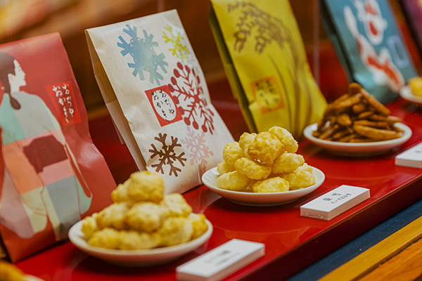 A close-up view of assorted Japanese snacks displayed in small bowls, with colorful packaging in the background on a red display tray.