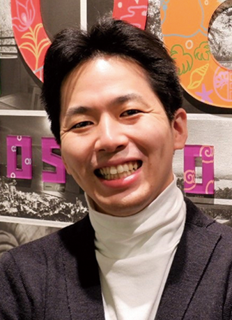 Portrait of a smiling person at Hoshino Resorts, wearing a dark jacket and white turtleneck, standing in front of a colorful background.