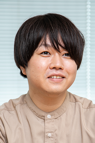 Portrait photo of a Drecom employee in a beige shirt, seated in front of window blinds, looking slightly to the side and smiling.