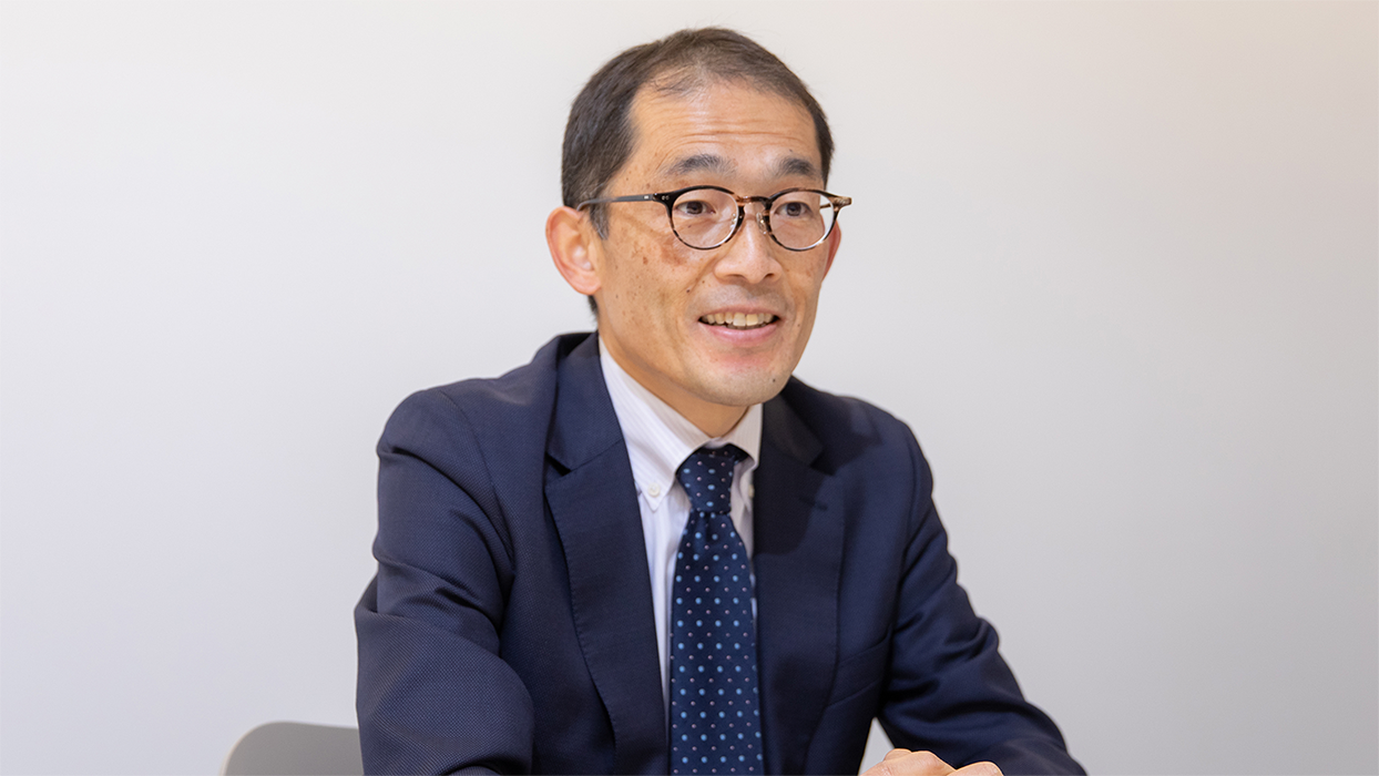 Business portrait of a person wearing a suit, seated and smiling, against a plain background. The person is wearing glasses and a polka dot tie.