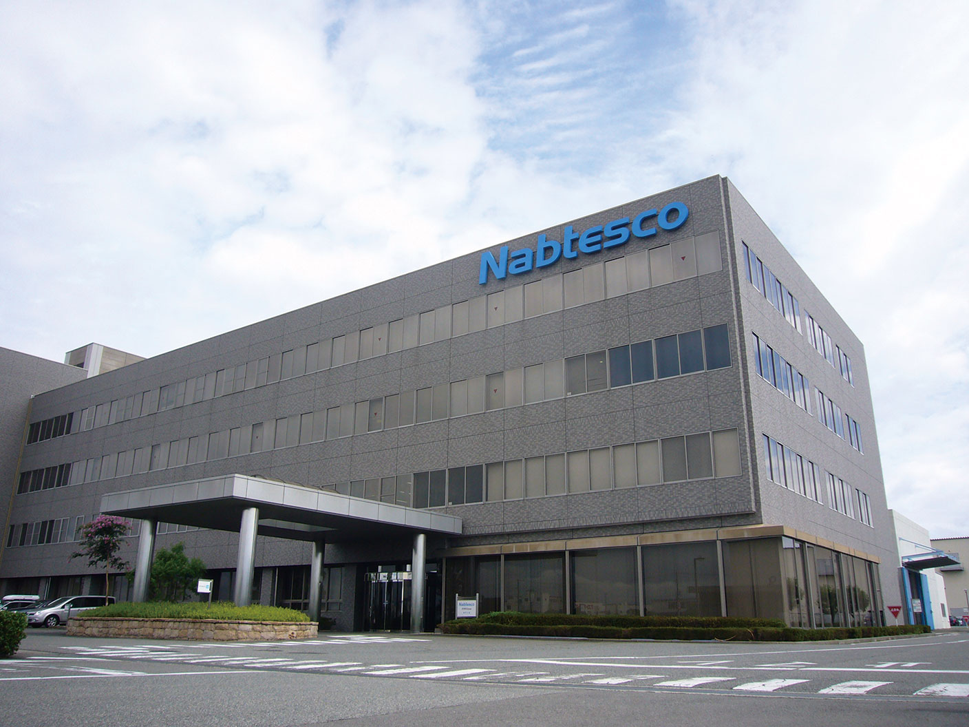 Exterior of a modern gray office building with "Nabtesco" written in blue letters on the facade.